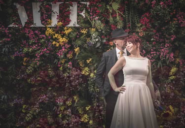 A smiling bride and groom pose in front of a colorful flower wall at a wedding venue.
