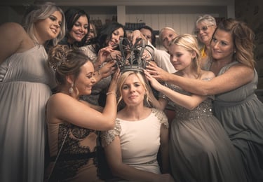 A group of bridesmaids and family placing a vintage metal crown on a bride's head at a wedding.