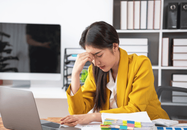 a woman in a yellow jacket is sitting at a desk with stressed face looking at laptop