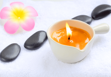 a massage candle in a cup container over a white cloth table with mindfulness stone and a flower