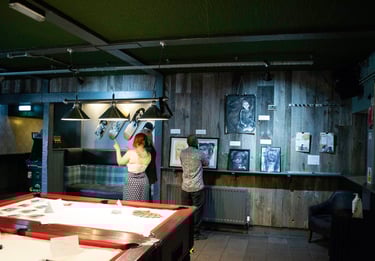 People at the exhibition looking and talking about art. Low lighting. Pool table in centre of room.