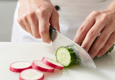 Crisp, sunlit kitchen action shot of a chef's hands slicing fresh cucumbers and radishes on a clean prep line, bright natural daylight.