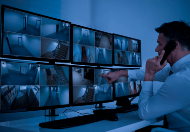 Security guard monitoring multiple CCTV camera feeds on a wall of video surveillance displays.