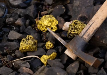 Natural raw gold nuggets lying on dark river rocks next to a vintage prospecting pickaxe.