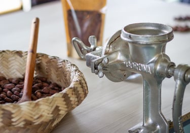 Vintage metal manual grinder next to a woven basket of raw cocoa beans for chocolate making.