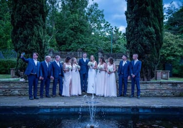 A wedding party in blue suits and pink bridesmaid dresses posing by an outdoor stone fountain.
