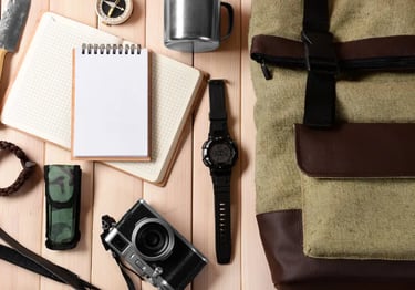 Survival gear and travel essentials including a canvas backpack, camera, notebook, and compass on wood.