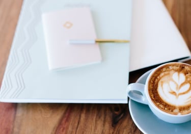 A cup of latte with foam art sits on a saucer next to a closed notebook with a pen on a wooden table