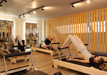Three people performing a bridge exercise on Pilates reformers in a well-lit studio with mirrors