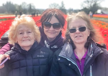 Three happy women wearing jackets and sunglasses smiling in a vibrant red tulip field.