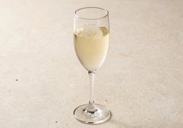 A crystal glass of sparkling wine with rising bubbles, set against a light beige stone surface. The lighting is bright and clean, typical of a refined Brazilian afternoon setting.