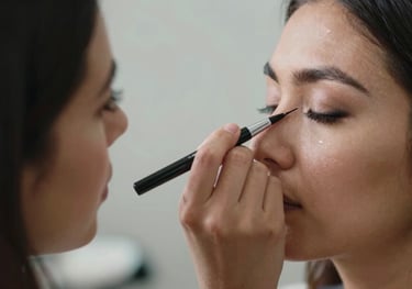 A close-up photograph of a professional makeup artist's hand precisely applying eyeliner on a client in a bright North American / NYC Hispanic studio.