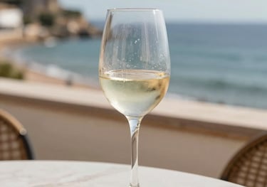 A chilled glass of white wine on a terrace overlooking a Spanish coastline. The lighting is bright and airy, capturing the condensation on the glass. Palette of sand and off-white.
