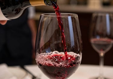 A close-up of a rich red wine being poured into a crystal glass. The setting is a refined restaurant in Southern Europe, with warm lighting and a hint of a professional kitchen in the background. Colors: deep burgundy and mahogany.
