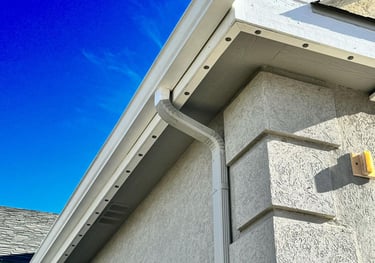 White aluminum seamless rain gutter and downspout installed on a stucco home exterior with a blue sky background.