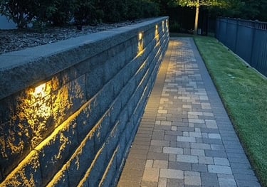 Illuminated stone paver walkway with landscape lighting integrated into a grey retaining wall.