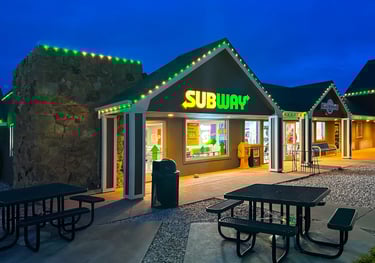 Subway sandwich shop exterior at night with green neon lights and outdoor patio seating.