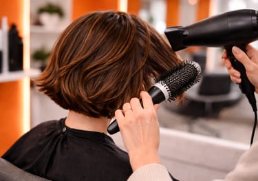 Professional hairstylist using a round brush and blow dryer to style a brunette bob haircut in a salon.