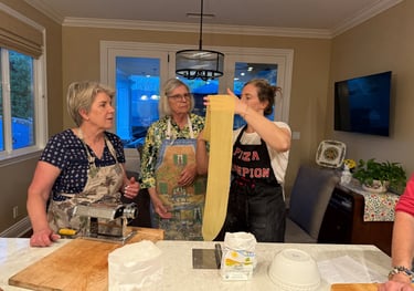 Three women wearing aprons make fresh pasta by hand using a stainless steel pasta roller machine in a home kitchen.