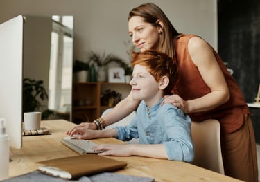 Lifestyle photo of woman standing behind son at desk in front of computer screen and keyboard.