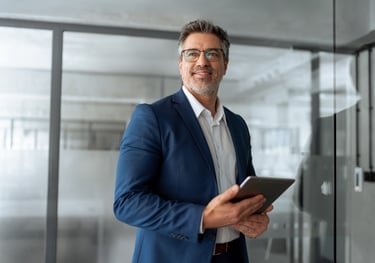 Smiling mature businessman in a blue suit holding a digital tablet in a modern corporate office.