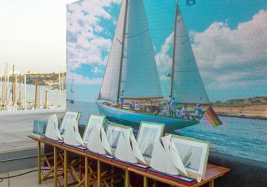 Pantalla led de fondo con imagen de barco navegando y delante mesa con trofeos y cuadros de fotos