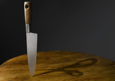 vertical kitchen knife balancing on its tip on a round wooden table with shadow of scissors