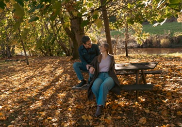 An engage couple sitting on a wooden picnic table in a park during autumn with golden fallen leaves.
