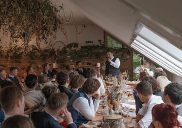 A best man in a kilt gives a wedding toast to guests at a rustic indoor reception venue.