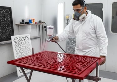 Professional painter in protective gear using a spray gun to apply red finish to an intricate laser-cut wood panel.
