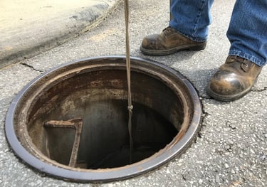 A persons workboots are visible as they stand next to an open manhole.