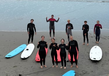 A group of surfers in wetsuits posing with surfboards on a sandy beach after a surfing lesson.