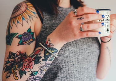 A woman with multiple tattoos, multiple rings and blue fingernails holds a coffee mug