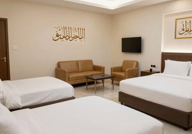 Interior of a spacious family suite in a hotel near the Haram. Multiple beds with crisp white linens, a seating area with tan leather chairs, and subtle gold-framed Arabic calligraphy on the cream wall.