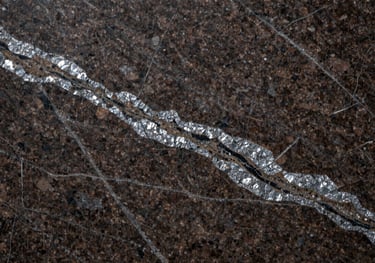 A close-up photograph of a polished dark brown granite slab with intricate silver and black mineral patterns, professional studio lighting, highlighting the high-quality finish and texture.