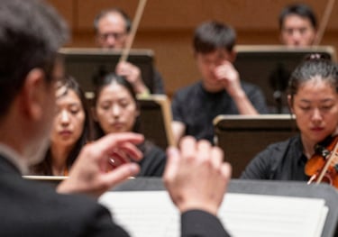 A photograph from behind a conductor's shoulder, showing their hands and the orchestra members looking up with focus during a rehearsal.