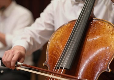 A close-up photograph of cello strings and a bow mid-motion. The focus is sharp on the resin dust and wood grain, with a background of other musicians in soft ice white shirts.