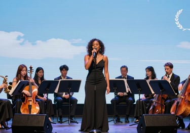 A vibrant photograph of a collaborative performance where an orchestral ensemble is backing a modern local jazz singer on a stage lit with steel blue and pale sky blue lights.