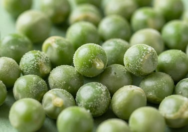 Close-up shot of freeze-dried green peas. The peas look crisp and vibrant green, set against a soft sage green surface.
