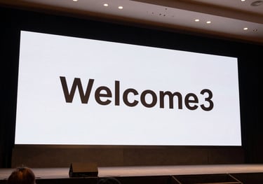 A wide shot of a corporate stage in a North American / US conference center. The screen displays 'Welcome' in a Montserrat font. The lighting uses warm bronze and dark charcoal tones.