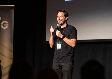 An MC on a stage at a North American / US tech conference. The lighting is professional with dark charcoal and muted gold stage accents. The MC is holding a microphone and smiling at the audience.