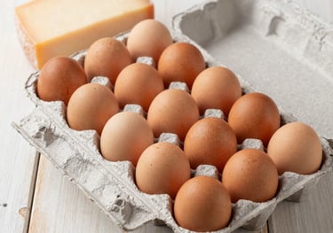A clean, bright photograph showing a dozen organic brown eggs in a pulp carton alongside a block of artisanal cheese on a rustic cream-colored wooden table.