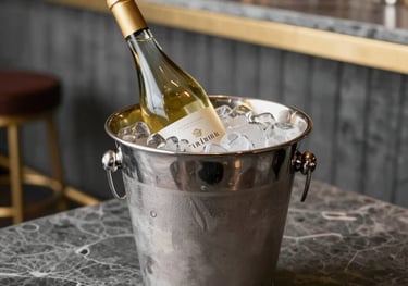 A bottle of white wine chilled in a silver bucket with ice, positioned on a slate tabletop. The setting is a trendy South American bar with elegant slate gray and gold decor.