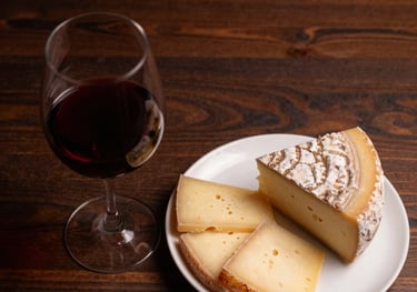 Top-down view of a glass of red wine next to a plate of artisanal Brazilian cheeses on a dark wood table. Warm, moody lighting highlights the deep red color of the wine.