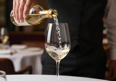 A waiter elegantly pouring white wine into a glass in a high-end Brazilian restaurant. The lighting is sophisticated, emphasizing the pale gold color of the wine.