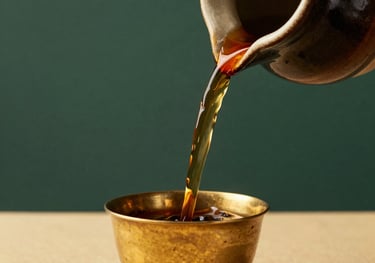 A close-up of a dark ceramic vessel pouring a rich, dark herbal liquid into a small brass cup, South Asian / Indian traditional medicine style, warm gold and deep green background.