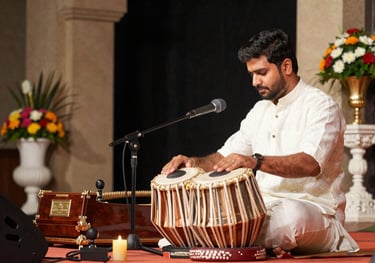 A professional stage setup for a Bhajan Sandhya featuring a tabla, harmonium, and elegant floral arrangements in a South Asian / Indian venue.