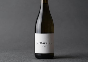 A minimalist studio shot of a Ceracore Syrah bottle. The label is clean and white with dark text. The lighting is moody and focused, highlighting the sleek curves of the glass. Tones of #2C3333 and #F7F7F7.
