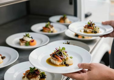 A close-up of beautifully plated gourmet meals being served from a professional mobile catering unit. International / Global cuisine. Natural, bright lighting. Colors: off-white and dark gray.