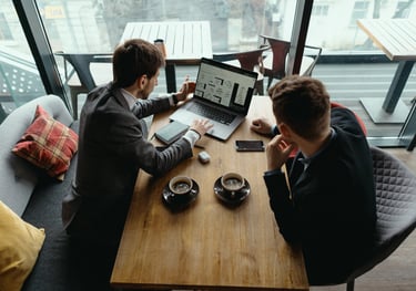Two businessmen in a cafe discussing data analytics on a laptop over coffee.